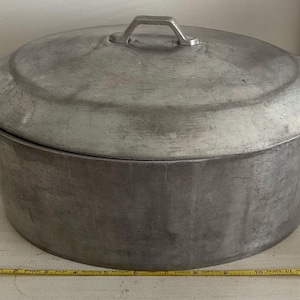 May include: A large, silver aluminum pot with a lid. The pot has a rounded bottom and a wide, flat rim. The lid has a handle on top.