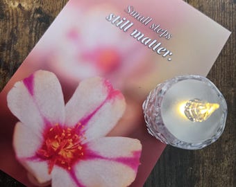 Könnte beinhalten: Eine Grußkarte mit einer rosa-weißen Blume und dem Text "Small steps still matter." Daneben befindet sich eine kleine, transparente LED-Kerze. Der Hintergrund ist eine dunkle Holzoberfläche.