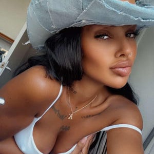 May include: A woman with dark hair is wearing a light blue denim cowboy hat with a brown leather patch. She is wearing a white tank top and has a gold necklace with a small pendant.