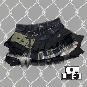 May include: A layered denim mini skirt with a patchwork design. Features include a black denim base, a pleated black skirt, and a green pocket with eyelets. Embellished with a cross pendant and a logo charm. The skirt has a distressed, punk aesthetic.