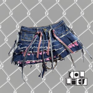 May include: A distressed denim skirt with frayed edges and pink fringe detailing. The skirt has decorative ribbons and the text "COL LIEU" in a white, stylized font. The skirt is set against a grey chain-link fence background.