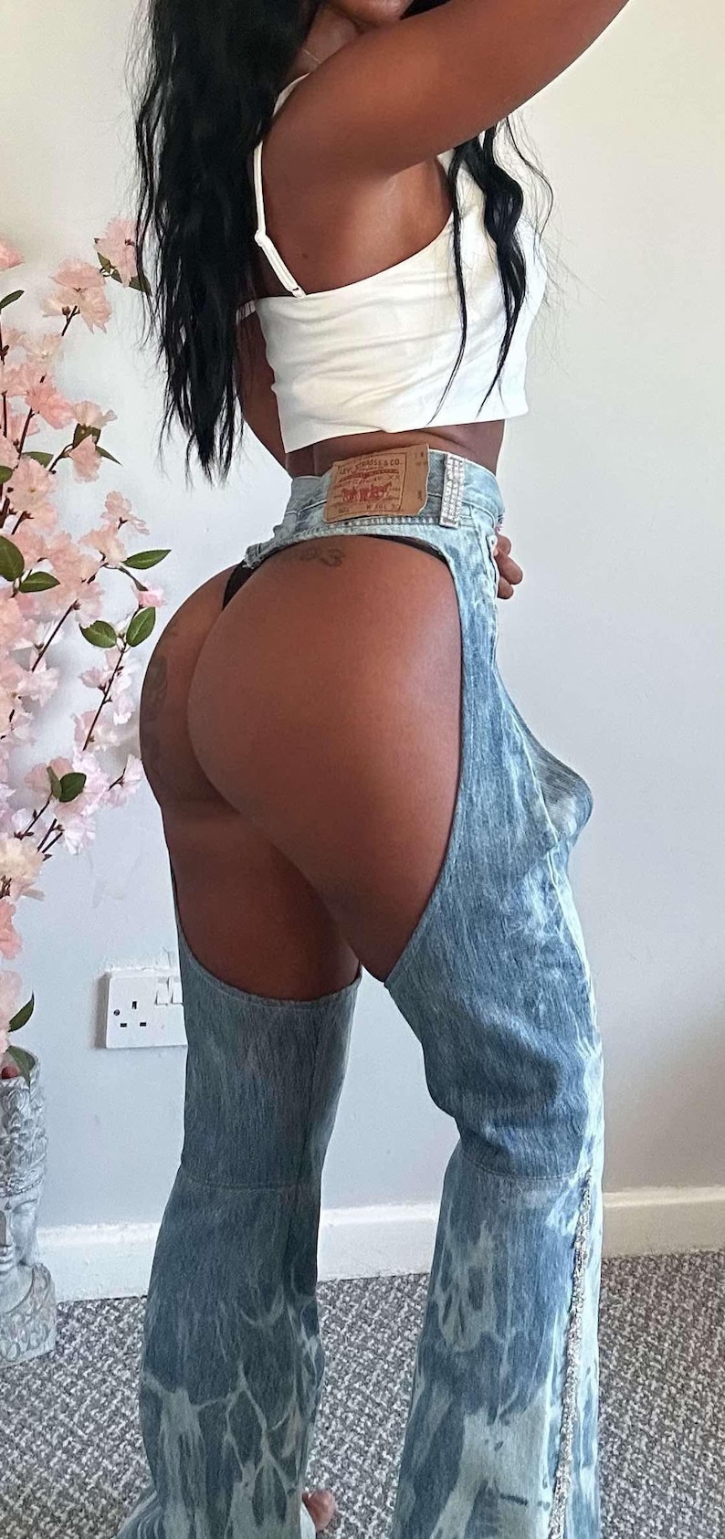 May include: A person wearing a white tank top and blue denim jeans with a cut-out on the side. The jeans have a light blue and white tie-dye pattern and a silver chain detail.