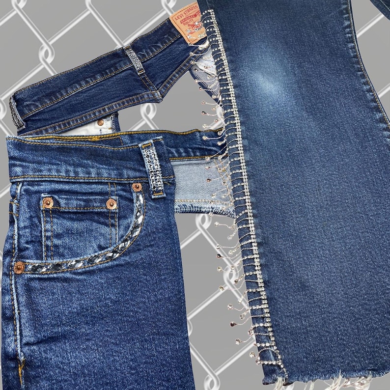 May include: Two pairs of blue denim jeans with rhinestones and a Levi's label. One pair has a rhinestone trim along the side seams and the other pair has a rhinestone trim around the pockets.