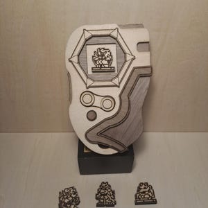 Puede incluir: Una réplica de madera de un dispositivo Digimon con una imagen pixelada de un Digimon en la pantalla. Tres pequeñas figuras de madera de Digimon están en primer plano.