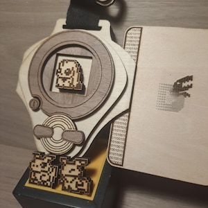 May include: A wooden, laser-cut replica of a digital pet device, with a small pixelated image of a creature inside the screen. The device has a strap and a binary code pattern on the side. Two smaller pixelated images of the creature are on a yellow base.