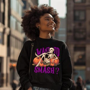 May include: Black sweatshirt with a Halloween-themed graphic. The design features a skeleton lounging on pumpkins with the text "WANT TO SMASH?" in purple. The sweatshirt has a crew neck and a relaxed fit.