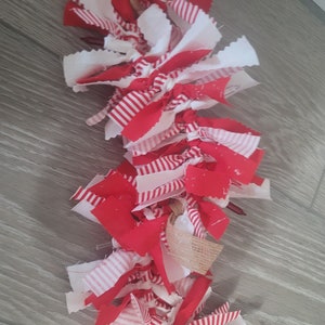May include: A red and white fabric garland with a burlap ribbon. The garland is made of strips of fabric that are tied together.