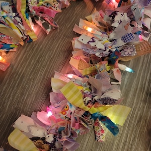 May include: A colorful fabric garland with white lights strung through it. The garland is made of various scraps of fabric in shades of pink, purple, blue, yellow, and green. The garland is perfect for adding a touch of whimsy to any room.