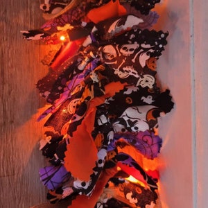 May include: A Halloween-themed fabric garland with orange, purple, and black fabric scraps. The garland is decorated with a variety of Halloween-themed patterns, including skulls, ghosts, and pumpkins. The garland is strung with white lights.