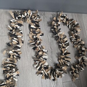 May include: Three decorative fabric garlands with a mix of black, white, and tan fabric strips. The garlands are arranged in a wavy pattern on a light gray wooden surface. A white electrical cord and plug are visible.