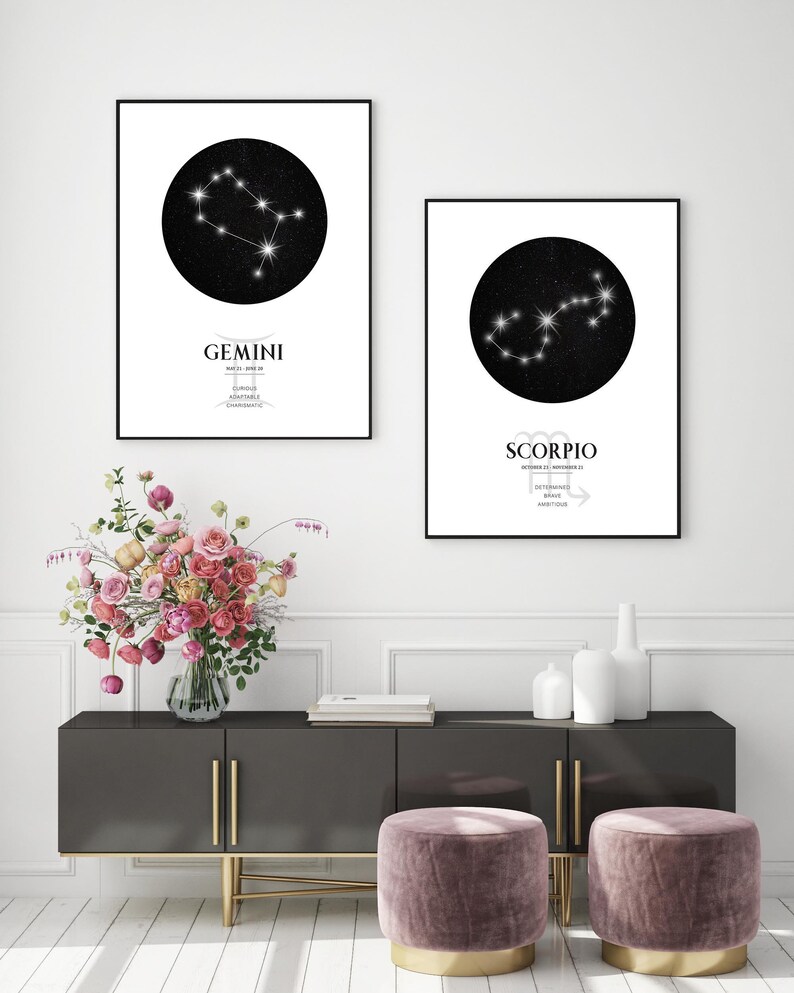 May include: Two black and white constellation prints with gold accents. The prints feature the zodiac signs Gemini and Scorpio with their corresponding dates and personality traits. The prints are hanging on a white wall above a dark grey dresser with gold accents. There are two pink velvet ottomans with gold accents in front of the dresser.