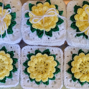 May include: A collection of handmade crochet squares, each featuring a yellow and green flower design on a white background. Some squares are tied with white string. The squares are ideal for various craft projects.