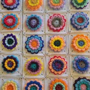 May include: A colorful crochet blanket featuring a repeating pattern of flower motifs. Each flower is a different color, including blue, orange, yellow, and pink, and is centered on a white square. The blanket is made of yarn and is likely handmade.