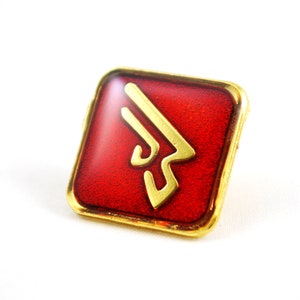 May include: A gold-colored pin with a red enamel background. The pin features a stylized, abstract design in gold.