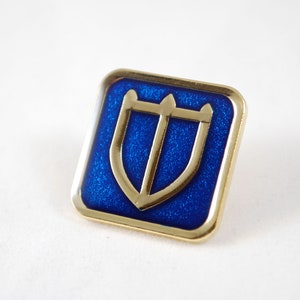 May include: A gold-colored pin with a blue enamel background. The pin features a stylized shield with three points at the top.