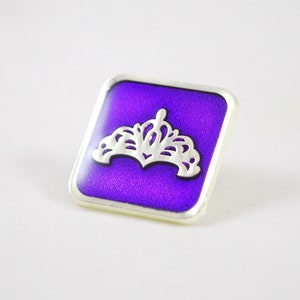 May include: A square-shaped pin with a silver border and a purple background. A silver crown design is centered on the pin.