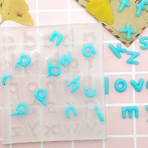 May include: A clear plastic mould with alphabet letters and a set of blue alphabet letter charms. The charms are scattered on a pink and white tiled surface.