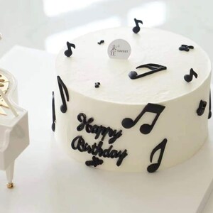 Puede incluir: Un pastel blanco decorado con notas musicales negras y las palabras "Happy Birthday".