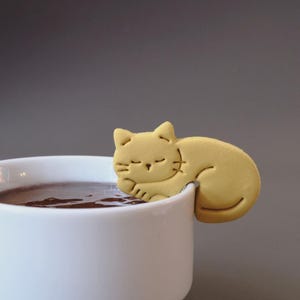 Könnte beinhalten: Eine matte, senfgelbe Keramikkatzenfigur, die so gestaltet ist, dass sie am Rand einer weißen Tasse eingehängt werden kann. Die Katze ist in einer Schlafposition mit geschlossenen Augen, kleinen Ohren und Schnurrhaaren dargestellt.