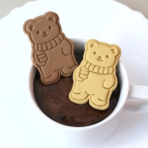 Könnte beinhalten: Zwei bärenförmige Kekse, einer hellbraun und einer beige, befinden sich in einer weißen Keramiktasse, die mit einer dunkelbraunen Flüssigkeit gefüllt ist. Jeder Keks zeigt ein geprägtes Bärenmotiv mit Schal sowie sichtbare Gesichtszüge und Pfoten.