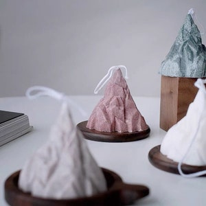 Peut inclure: Des bougies en forme de montagne dans les tons rose, blanc et vert, chacune avec une mèche en ficelle blanche. Les bougies sont posées sur de petits socles ronds en bois foncé. Le fond est blanc neutre.