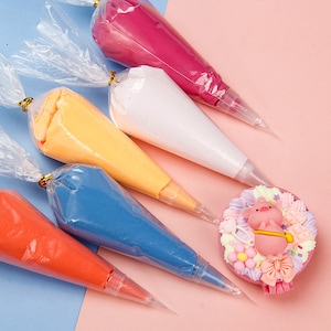 Könnte beinhalten: Fünf Spritzbeutel gefüllt mit unterschiedlich farbigem Frosting, darunter rosa, weiß, gelb, blau und orange. Die Beutel sind fächerförmig auf einer rosa und blauen Oberfläche angeordnet. Ein kleiner rosa Tortendekor mit einem Schweinemotiv befindet sich in der Mitte des Bildes.