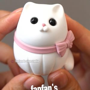 May include: A small white clay cat figurine with black eyes, a pink nose, and a pink collar featuring a bow on the side. The text "Fan" is visible at the top, "@fanfandesignstudio" is on the left, and "fanfan's Clay Crafts" is at the bottom.