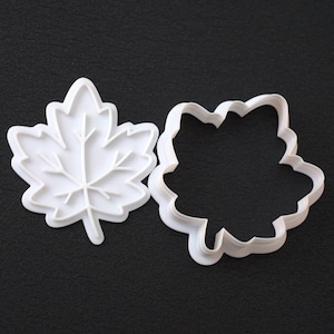 Puede incluir: Un juego de dos cortadores de galletas de plástico blanco con forma de hoja de arce. Una pieza es un cortador de contorno sólido y la otra es un sello detallado que muestra las venas internas de la hoja para crear un diseño en relieve.