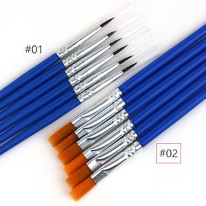 Könnte beinhalten: Ein Set von Pinseln mit blauen Griffen und silbernen Zwingen. Die mit #01 gekennzeichnete Gruppe hat feine, spitz zulaufende schwarze Borsten mit durchsichtigen Schutzkappen. Die Gruppe #02 hat flache, goldbraune synthetische Borsten.