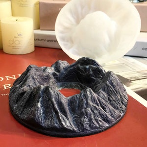 Puede incluir: Un porta incienso gris en forma de montaña con una abertura circular en el centro. El porta incienso está hecho de un material texturizado y tiene un color gris oscuro.