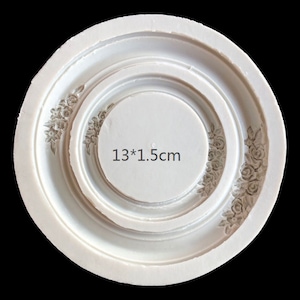 Könnte beinhalten: Weiße Silikonform mit einem runden Design mit einem floralen Muster. Die Form hat einen Durchmesser von 13 cm und eine Tiefe von 1,5 cm.