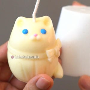 Könnte beinhalten: Eine blassgelbe, katzenförmige Kerze mit weißem Docht, blauen Augen und einer rosa Nase. Die Katzenfigur hat eine Schleife an der Seite und den Text fanfandesignstudio auf dem Körper. Daneben befindet sich eine schlichte, weiße zylindrische Form.