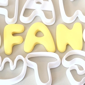 Könnte beinhalten: Eine Sammlung weißer Kunststoff-Ausstechformen in Form von Buchstaben des Alphabets, angeordnet neben gelben Fondant- oder Teigausstichen, die die Buchstaben F, A und N in einer runden, fetten serifenlosen Schriftart bilden.