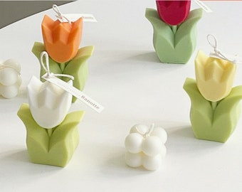 May include: Assortment of colorful tulip-shaped candles with green leaves. The candles are arranged on a white surface.