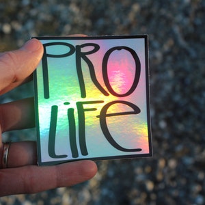 May include: A square sticker with a holographic finish. The sticker has black text that reads "PRO LIFE" in a handwritten style.