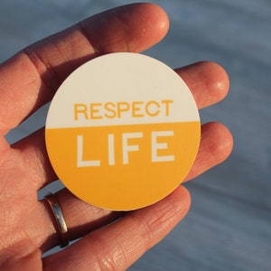 May include: A round sticker with a white top half and a yellow bottom half. The words "RESPECT LIFE" are printed in white on the sticker.