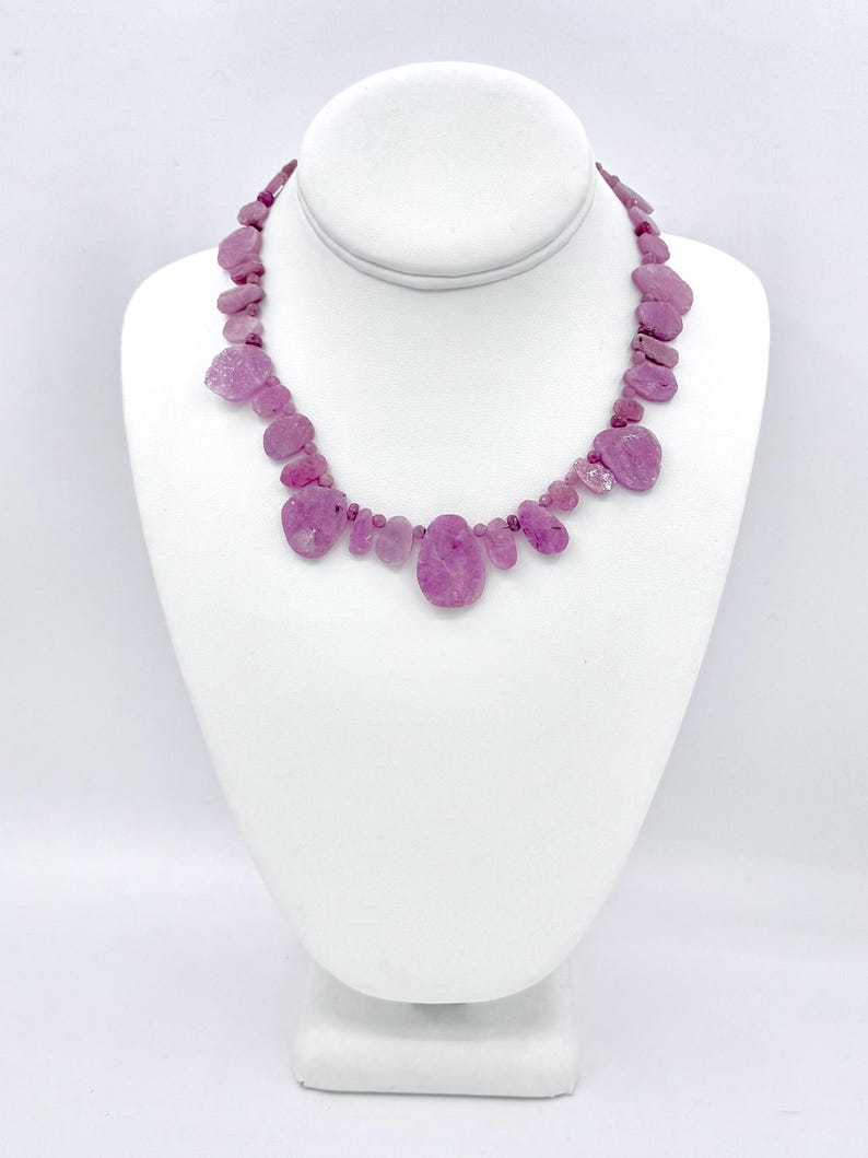 May include: A pink gemstone necklace displayed on a white jewelry bust. The necklace features irregularly shaped, polished pink stones of varying sizes, strung together to form a statement piece. The stones range in color from light to deep pink.