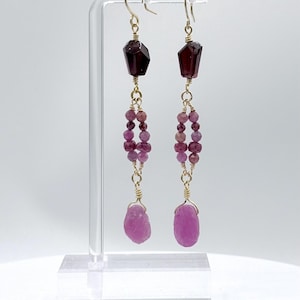 May include: Dangle earrings with dark red faceted gemstones, connected by gold wire to a strand of small, pink beads. The earrings end with a large, irregular pink gemstone. The earrings are displayed on a clear stand.