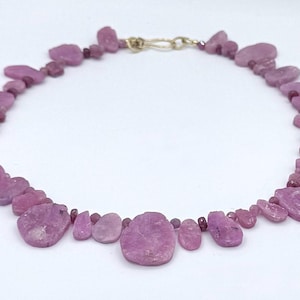 May include: A necklace of irregular, pale purple gemstones. The stones are linked by small, round beads and a gold-coloured clasp. The necklace is shown against a white background, emphasising the colour and texture of the stones.