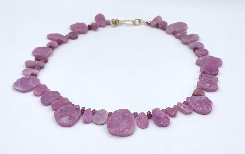 May include: A necklace featuring irregular, light purple gemstones. The stones are connected by small, round beads and a gold-colored clasp. The necklace is displayed on a white background, highlighting the color and texture of the stones.