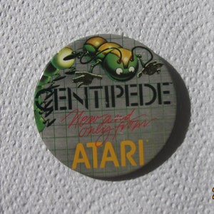 May include: A round, silver pin with a green and yellow centipede graphic. The pin says "CENTIPEDE" in red letters and "ATARI" in black letters.
