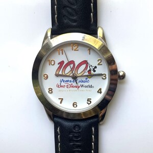 May include: A black leather wristwatch with a white face and gold trim. The face features the text "100 Years of Magic Walt Disney World" and a cartoon image of Mickey Mouse.