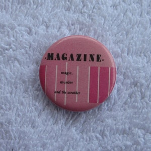 May include: A pink and white button badge with the text "MAGAZINE." and the words "magic, murder and the weather" printed on it.