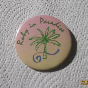 May include: A pink and yellow round button with the text "Ruby in Paradise" and a green palm tree with a blue wave.