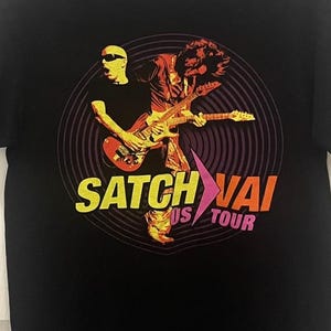 May include: Black t-shirt featuring a graphic of a musician playing a guitar, with the text "Satch Vai US Tour" in yellow and orange. The design is set within a circular pattern.