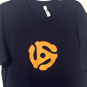 May include: Navy blue t-shirt with a yellow and orange 45 RPM record adapter graphic. The short-sleeved shirt is hanging on a white door. The graphic is centered on the front of the shirt.