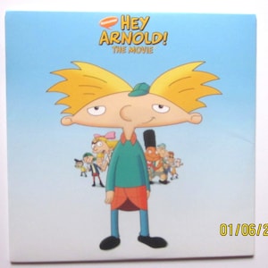 May include: A cartoon illustration of Arnold from the Hey Arnold! animated television series. Arnold is wearing a green shirt, red pants, and a green hat. He is standing in front of a blue background with other characters from the show behind him. The text "Hey Arnold! The Movie" is at the top of the image.
