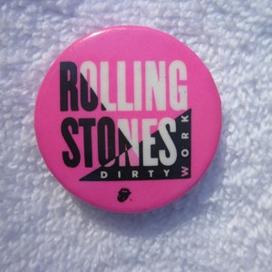 May include: A round, pink button with the words "Rolling Stones" in white and black, and the words "Dirty Work" in black. The Rolling Stones' iconic tongue logo is at the bottom.