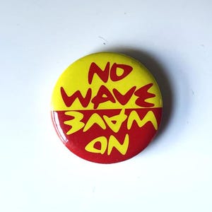 May include: A red and yellow button with the text "NO WAVE" on the top half and "NO WAVE" on the bottom half, with the text reversed on each half.