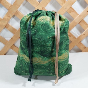 May include: A green drawstring bag with a swirling gold and green pattern. The bag has a dark green drawstring on the left and a tan drawstring on the right. The bag is made of fabric and is sitting on a white surface.